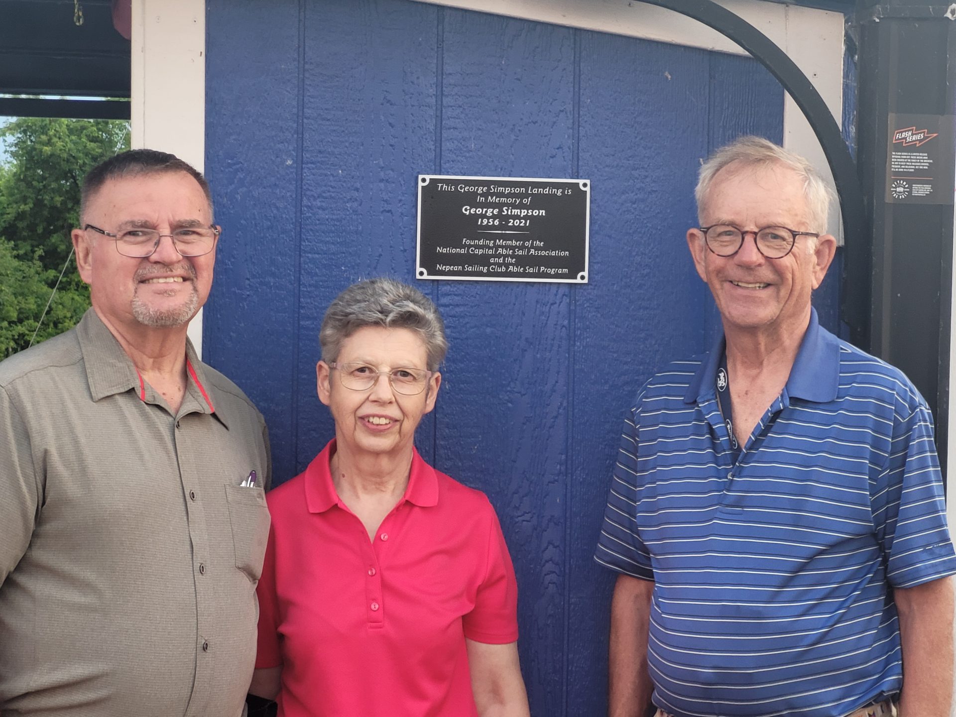 Three people standing before a plaque dedication to George Simpson
