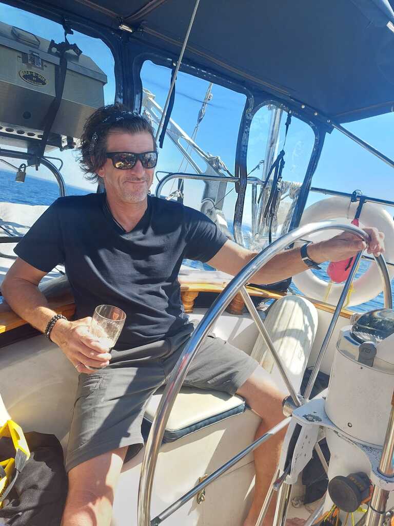 person holding a glass in one hand while steering a sailboat with steering wheel in the other